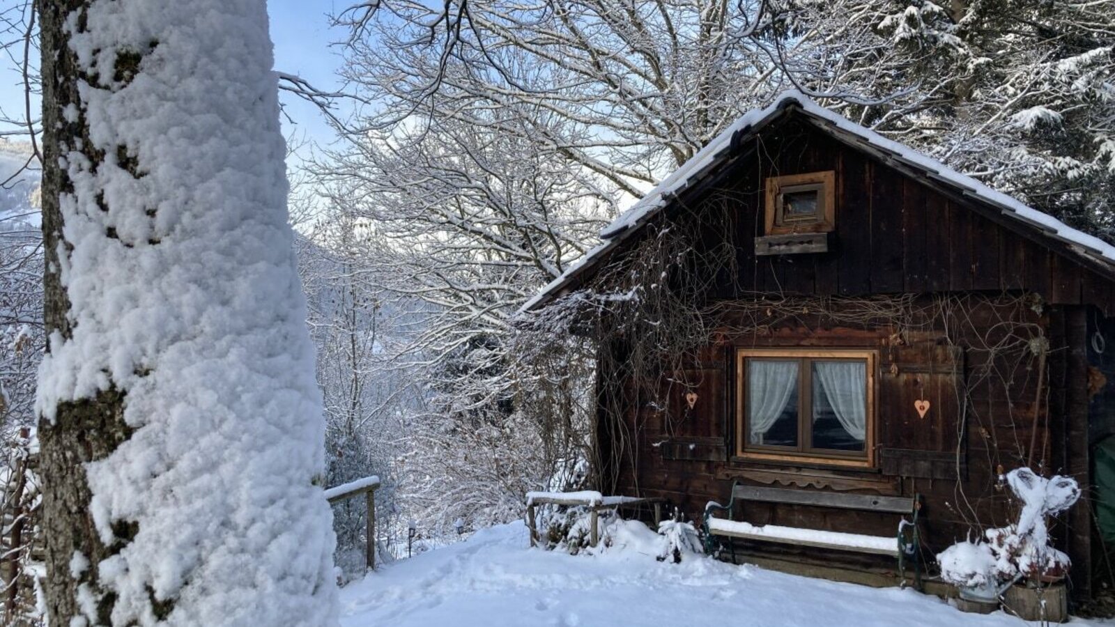 The snowy winter landscape surrounds the wooden cabin with its window beams, the red heart and the cozy bench in front of it.