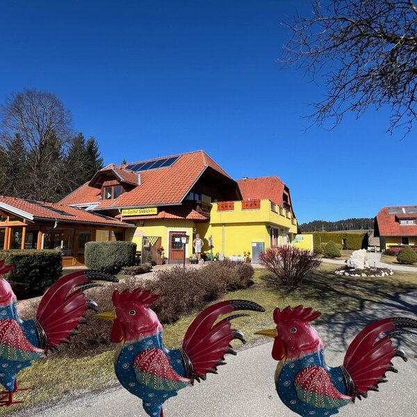 Exterior view of the farmhouse with a yellow building, red roof, solar panels, and surrounding green area under a blue sky.