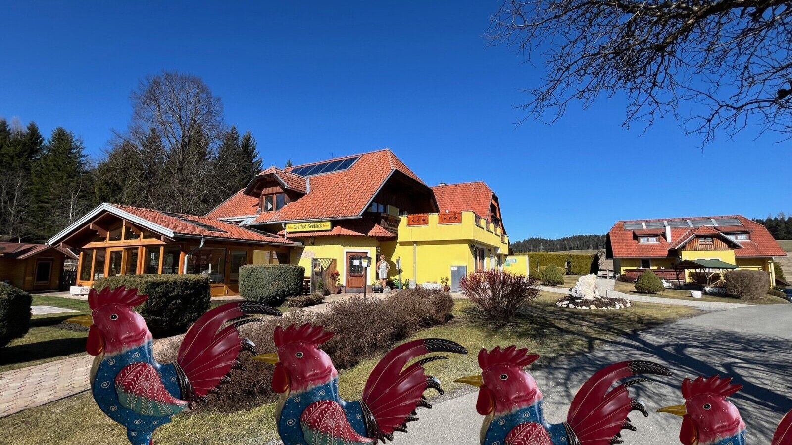 Exterior view of the farmhouse with a yellow building, red roof, solar panels, and surrounding green area under a blue sky.