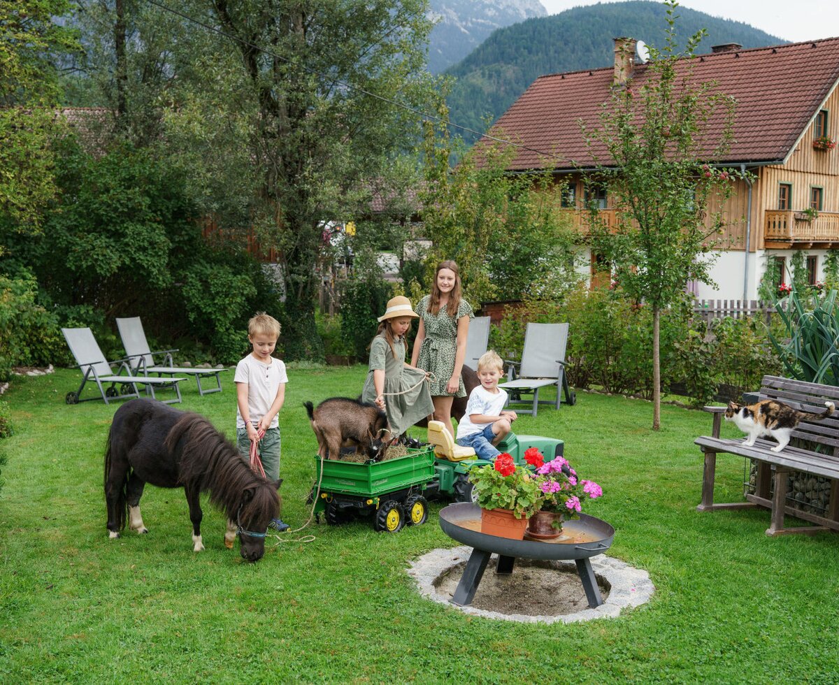 Children playing with a pony and a goat in the Bauernhof garden, featuring a toy tractor and a cat on a bench.