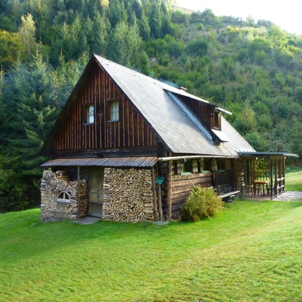 The wooden farmhouse, set against a forested hillside, features a covered porch with outdoor seating and a section of stacked firewood.
