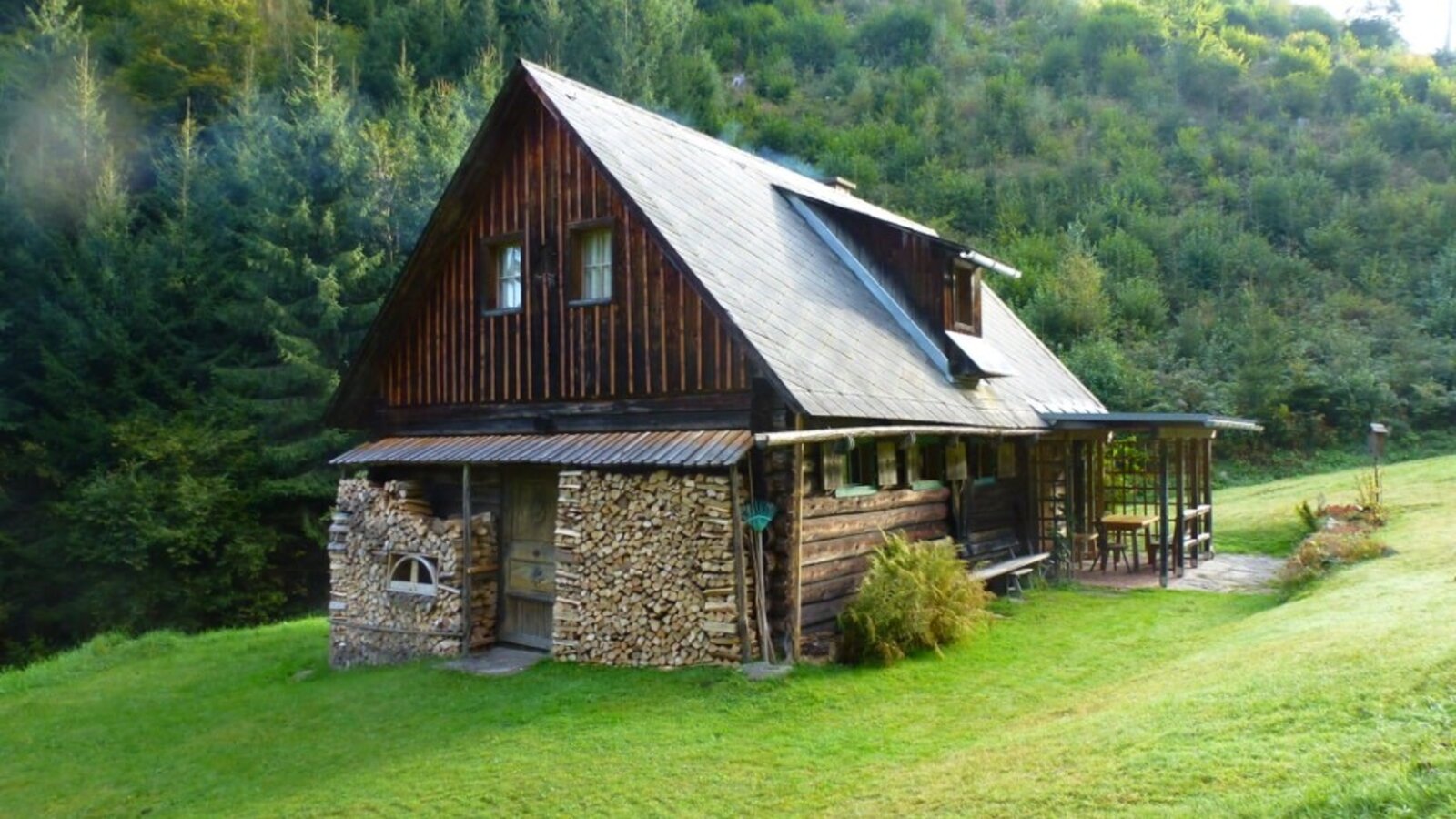 The wooden farmhouse, set against a forested hillside, features a covered porch with outdoor seating and a section of stacked firewood.