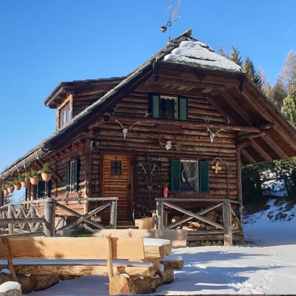 The exterior of the farmhouse with a rustic wooden bench on snow-covered ground, surrounded by pine trees.