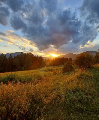 Sunset over the natural landscape surrounding the Farm House, featuring fields, forests, and distant mountains under a cloudy sky.