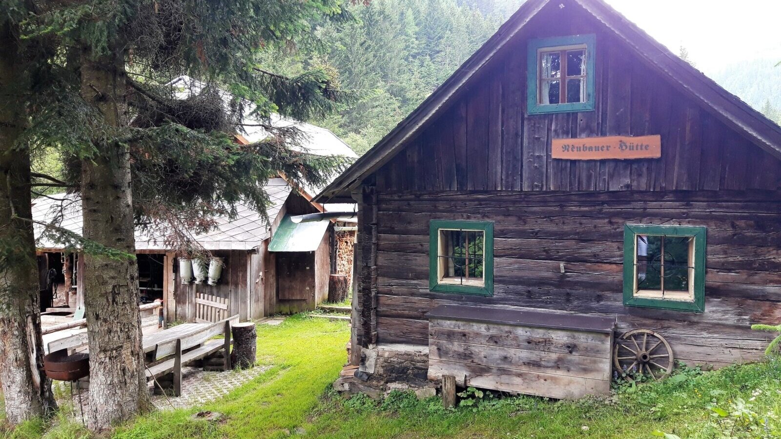 The exterior of the farmhouse features a dark wooden main building with green-framed windows and a "Neubauer Hütte" sign, situated in a mountainous, forested landscape with an adjacent outdoor seating area.