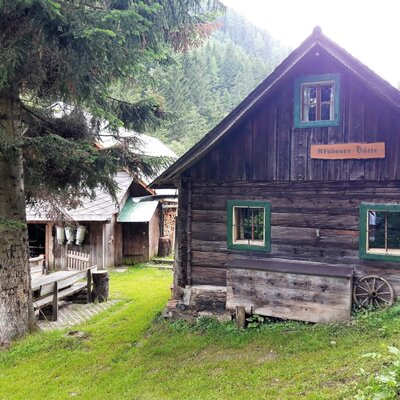 The exterior of the farmhouse features a dark wooden main building with green-framed windows and a "Neubauer Hütte" sign, situated in a mountainous, forested landscape with an adjacent outdoor seating area.