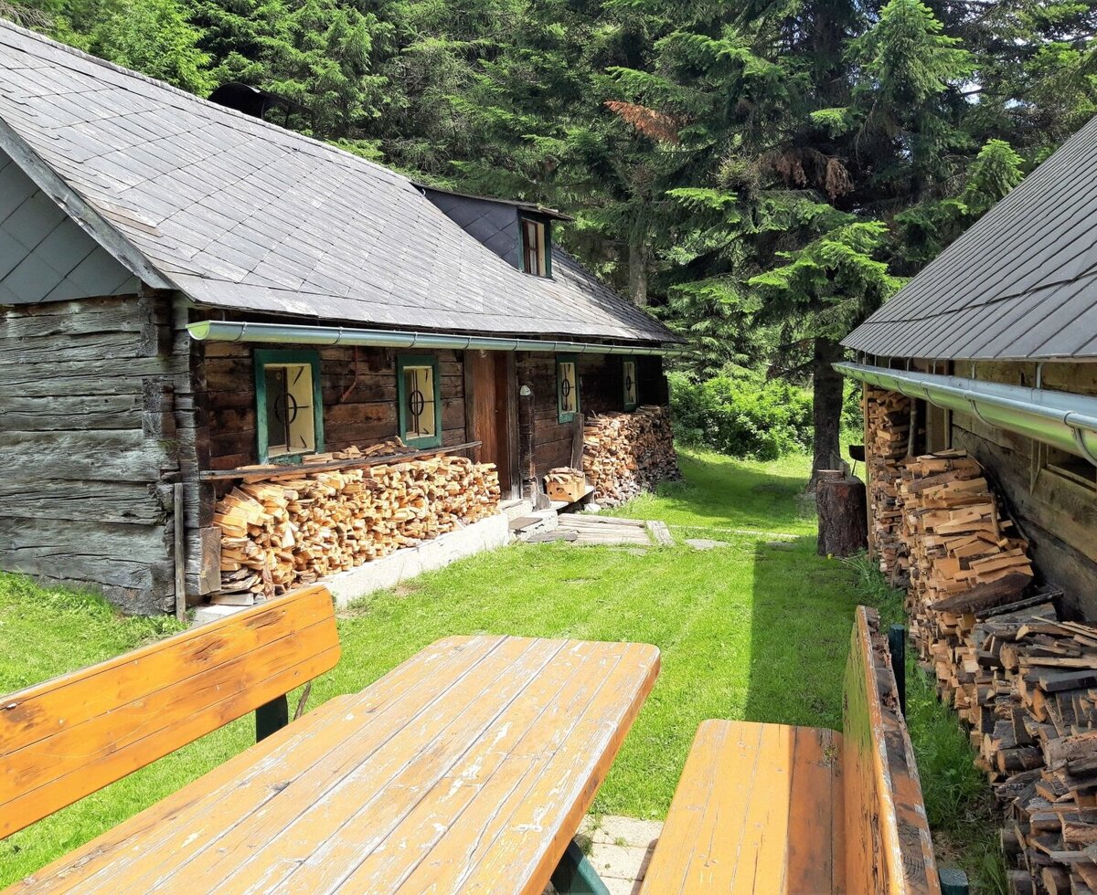 The farmhouse exterior features two wooden buildings, extensive stacked firewood, and an outdoor picnic table with benches on a grassy lawn.