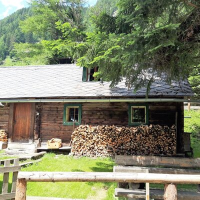 The wooden exterior of the farmhouse with a grey roof, stacked firewood, and surrounding trees.