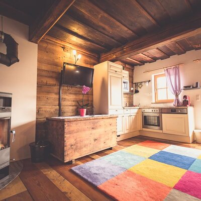 A cozy living area in the Ottls Hütte featuring a warm wood-burning stove, rustic timber beams, and an open-plan kitchen.