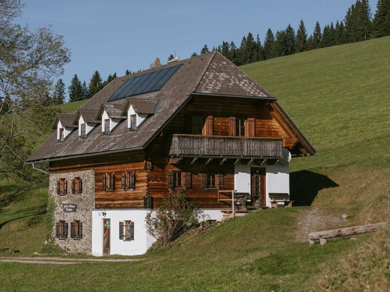 Exterior view of the farmhouse, featuring a wood and stone facade, a balcony, and solar panels on the roof.