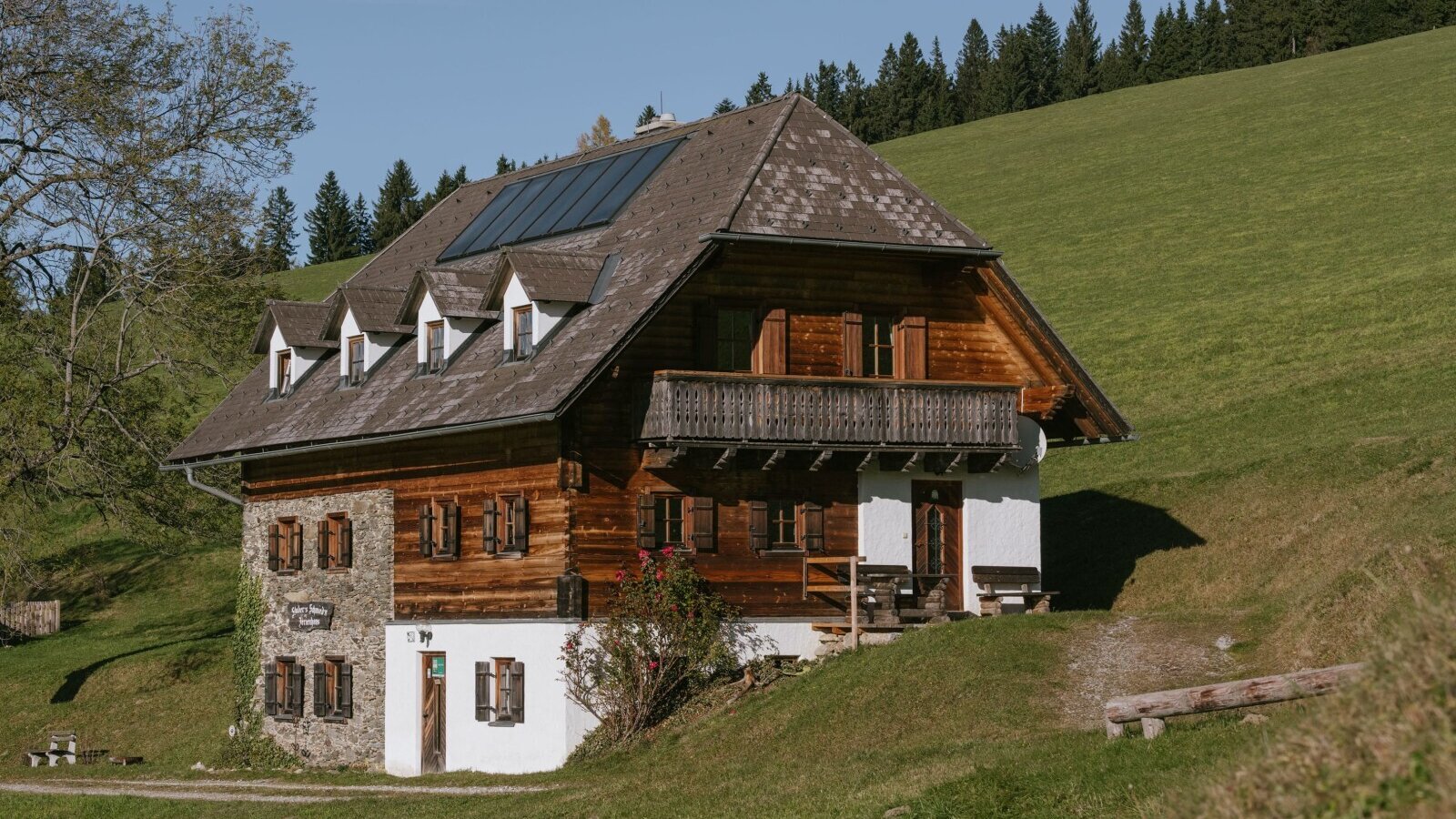 Exterior view of the farmhouse, featuring a wood and stone facade, a balcony, and solar panels on the roof.
