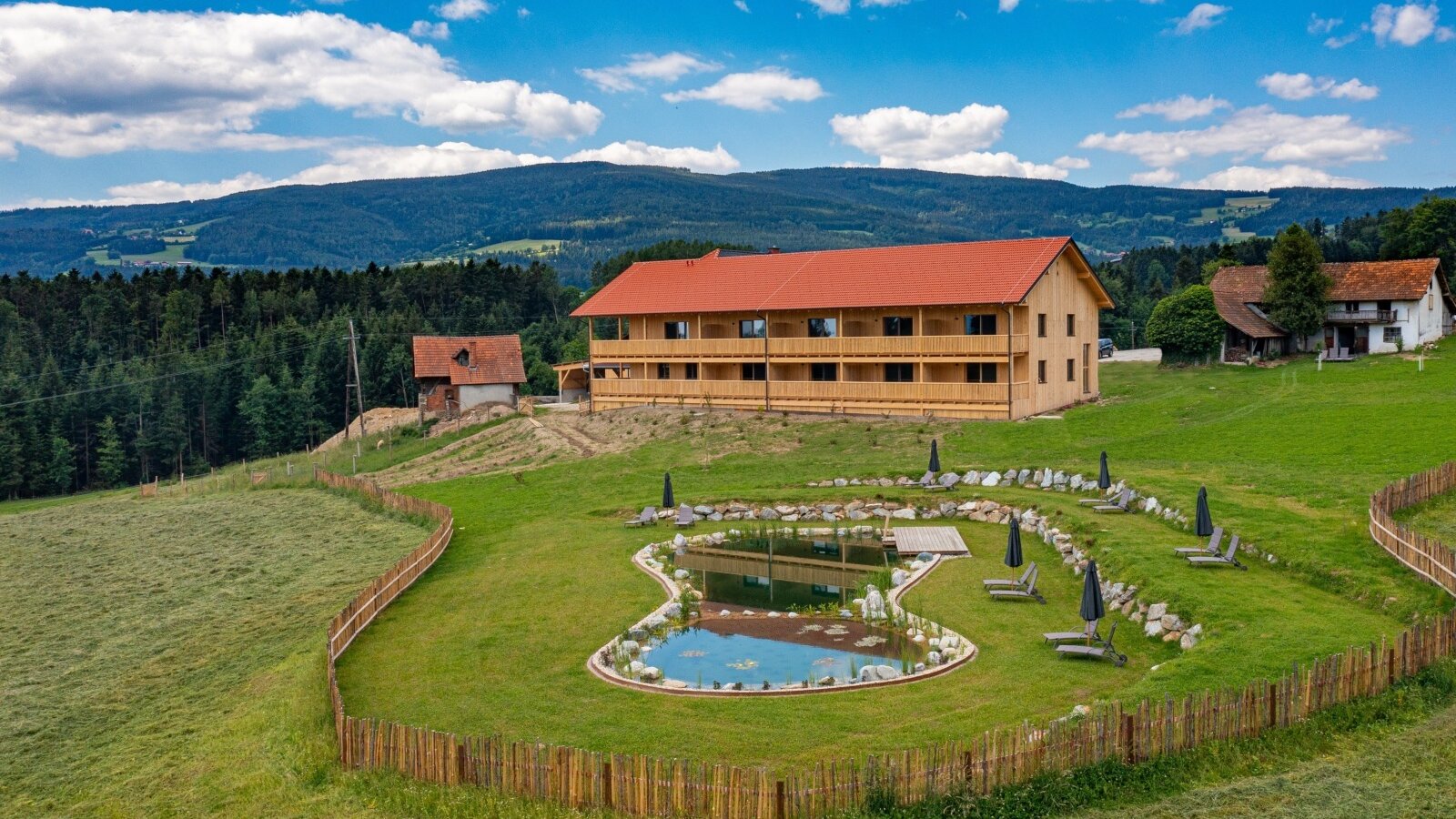 The new annexe of the Farm House, featuring private balconies, overlooks a pond with sun loungers, surrounded by green hills and distant mountains.