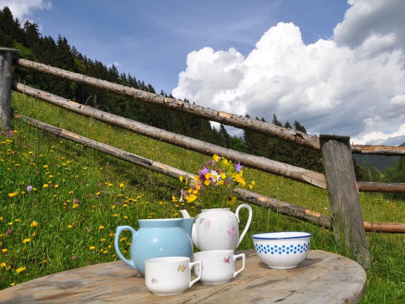 A seating area at the farmhouse, featuring a wooden table with a tea set and wildflowers, overlooking a fenced hillside.