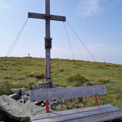 A wooden summit cross and a bench on a grassy mountain peak.