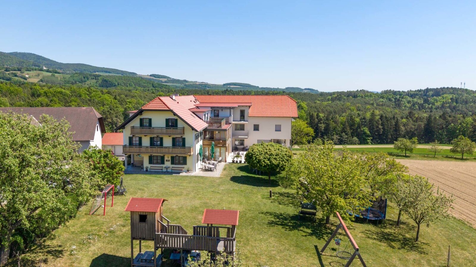 The farmhouse with balconies and an outdoor terrace, surrounded by a garden with a children's playground and the landscape of Pöllauer Tal.