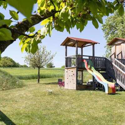 Children's playground at the Bauernhof, featuring a wooden play tower, a colorful slide, and a climbing wall, surrounded by green fields.