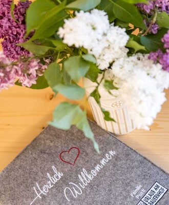Felt mat with 'Herzlich Willkommen' (Welcome) and the 'Urlaub am Bauernhof' (Holiday on the farm) logo, alongside a bouquet of lilacs on a wooden table.