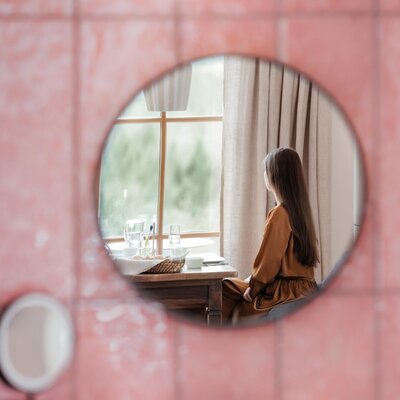 A round mirror on a tiled wall reflects a farmhouse room, featuring a window table overlooking nature.