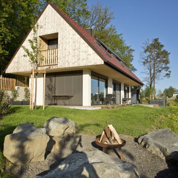 The farm house exterior features an A-frame roof with an upper balcony, a ground-floor patio, and a fire pit surrounded by rocks in the garden.