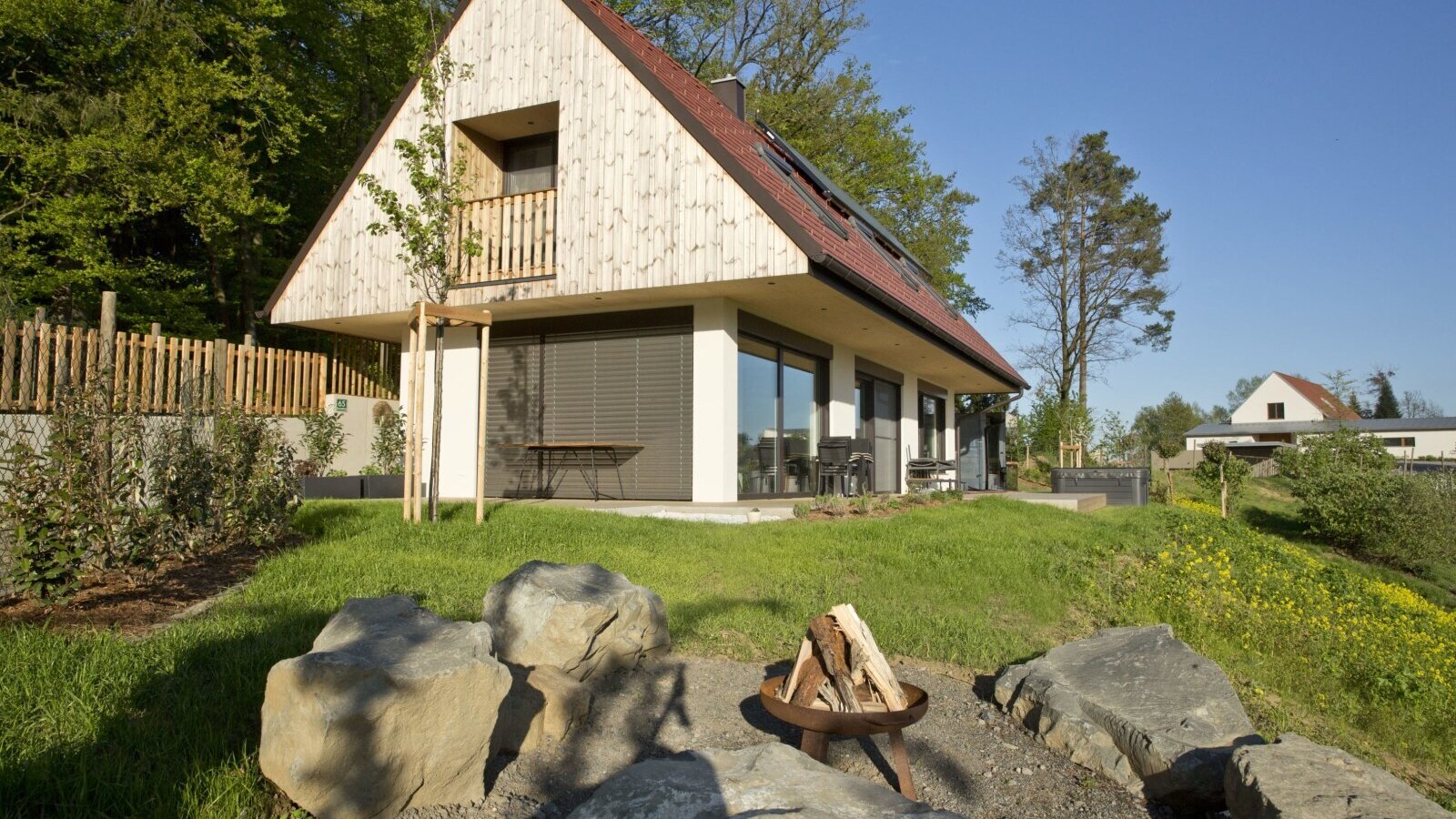 The farm house exterior features an A-frame roof with an upper balcony, a ground-floor patio, and a fire pit surrounded by rocks in the garden.