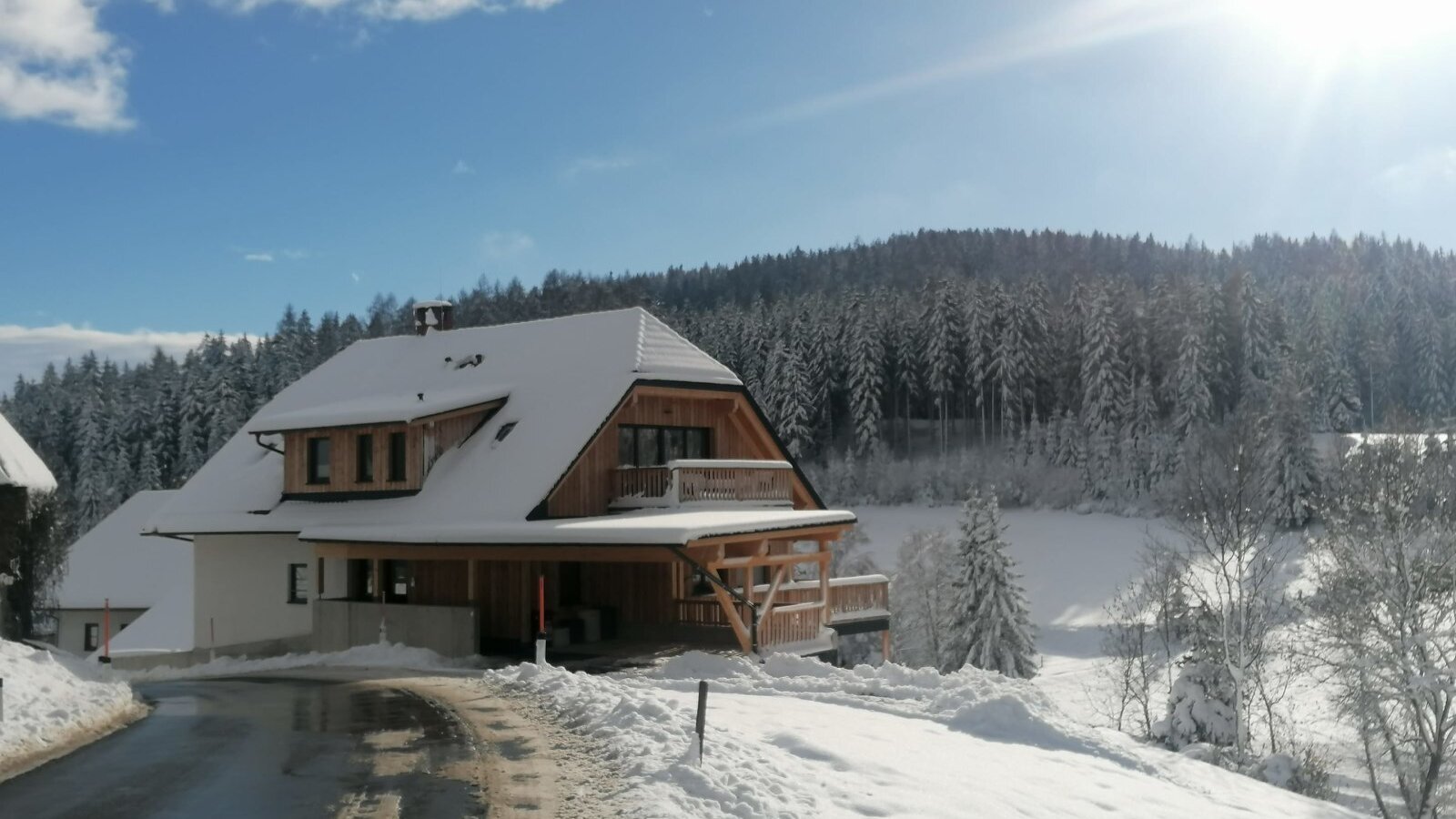 The Farm House exterior in winter, featuring a snow-covered roof, wooden elements, and a surrounding forest landscape, with a cleared access road.
