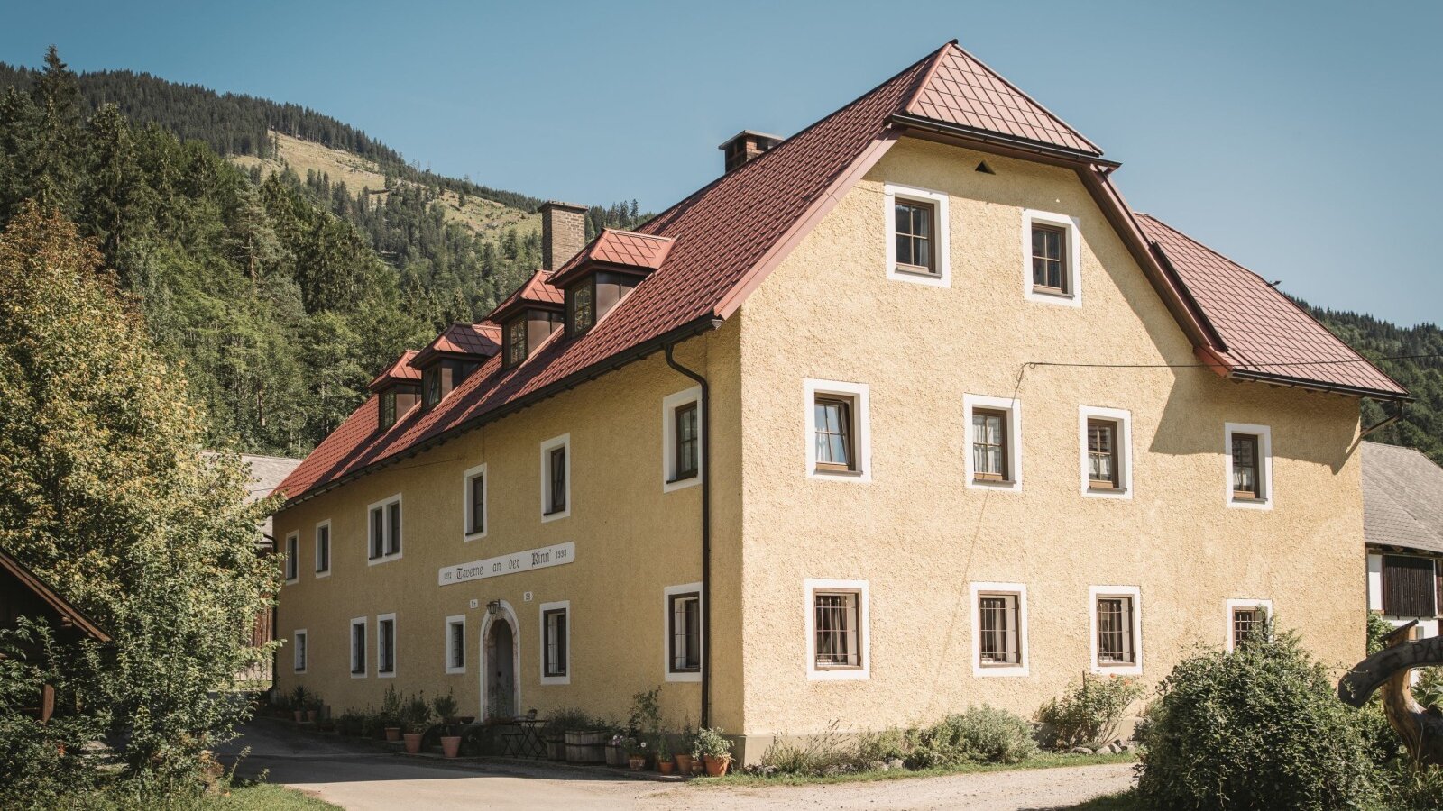The yellow-painted farm house exterior with a red tiled roof, located by a dirt path and backed by green forested hills.