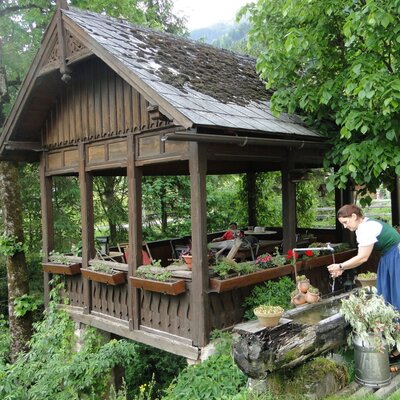 The farmhouse features a traditional wooden gazebo with outdoor dining seating and flower boxes, complemented by a rustic log water fountain.