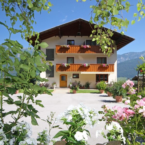 The exterior of the Farm House, featuring balconies with flower boxes, a paved courtyard, a surrounding garden with various flowers, and distant mountains.