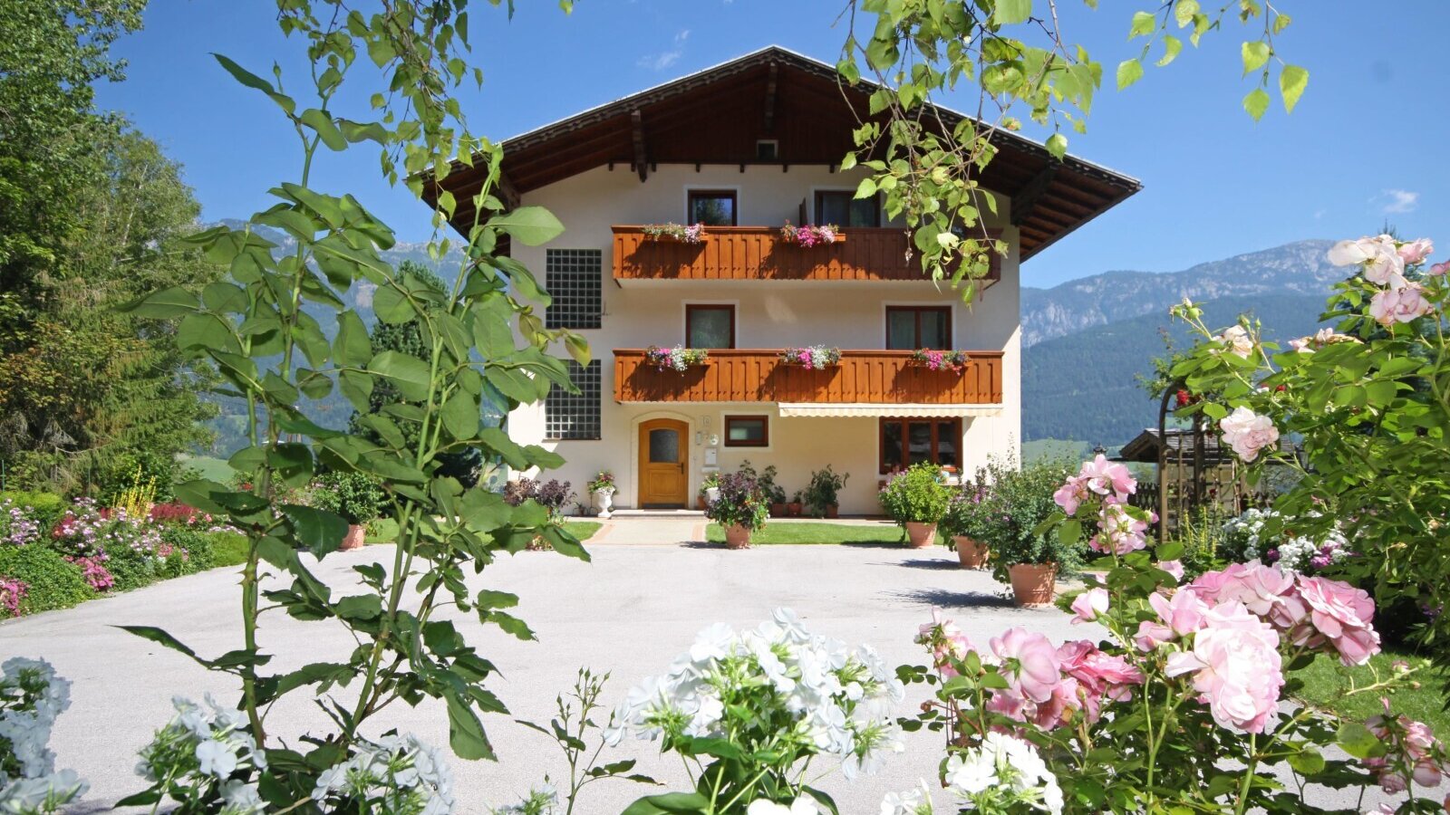 The exterior of the Farm House, featuring balconies with flower boxes, a paved courtyard, a surrounding garden with various flowers, and distant mountains.