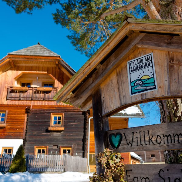 The exterior of the farmhouse during winter, featuring a traditional wooden welcome sign and snow on the ground.