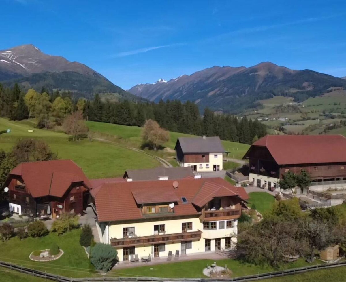 The farmhouse with its buildings, surrounded by green meadows and forests, with a mountain range in the background.