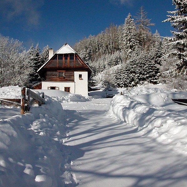 The farmhouse exterior in winter, with deep snow covering the ground and surrounding trees, and a snow-bordered path leading towards the building.