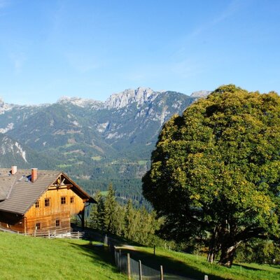 The wooden farmhouse is situated on a grassy slope with a view of the mountains.