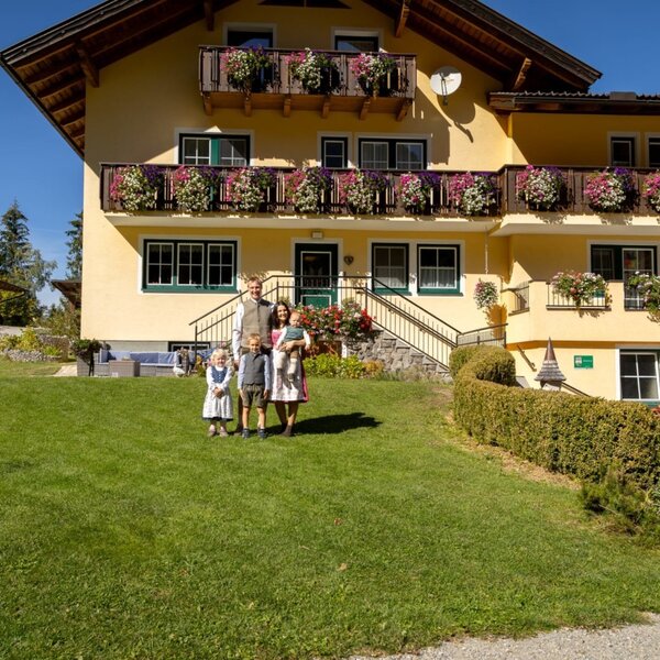 The inviting farmhouse featuring a well-kept green lawn, flower-filled balconies, and a welcoming atmosphere under a clear blue sky.