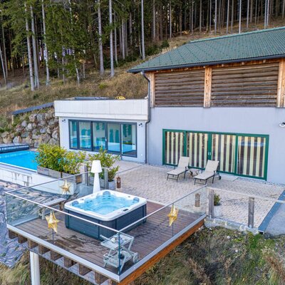 The holiday home's inviting terrace featuring a modern hot tub on a wooden deck, an outdoor pool, and comfortable loungers set against a forest backdrop.