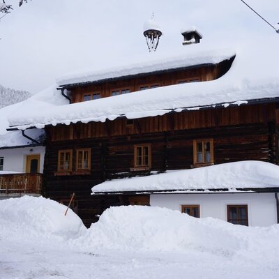 Farm house exterior in winter, featuring a wooden facade, snow-covered roof, and deep snowdrifts around the property.