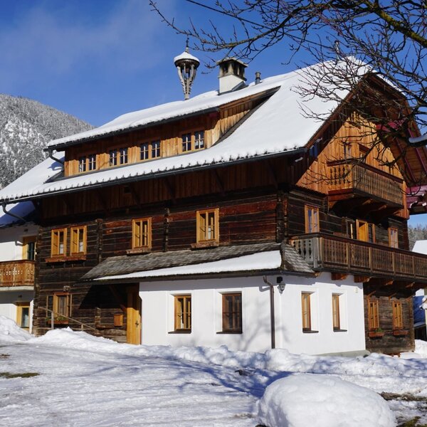 The Farm House features traditional wooden construction with white walls, multiple balconies, and a snow-covered roof, surrounded by winter scenery and mountains.