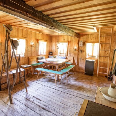 The main living area of the farmhouse features a wooden dining table with benches, an all-wood interior, and a ladder providing access to an upper level.
