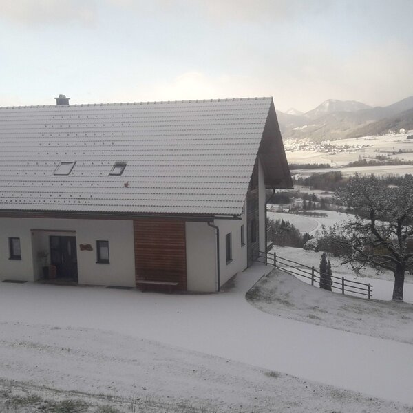 The snow-covered farmhouse, with its white exterior and gabled roof, overlooks a winter valley landscape with distant mountains.