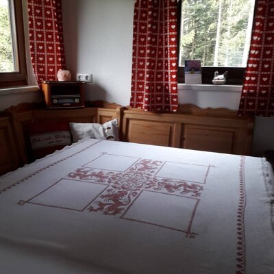 The farmhouse's traditional dining area offers wooden bench seating and a large table with an embroidered tablecloth, with views of trees from the windows.