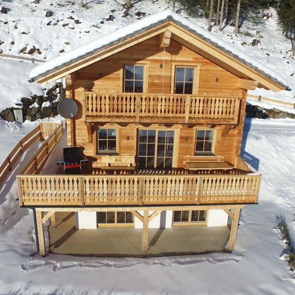 The wooden farmhouse with two levels of balconies is set amidst a snowy, tree-lined landscape.