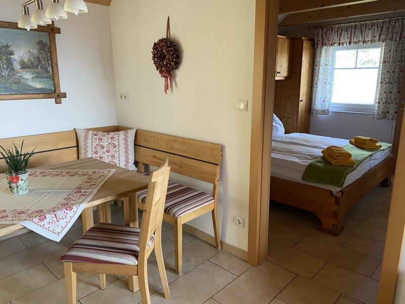 The dining area of the Farm House, equipped with solid wood bench seating and a table, leading into a bedroom furnished with a wooden bed.
