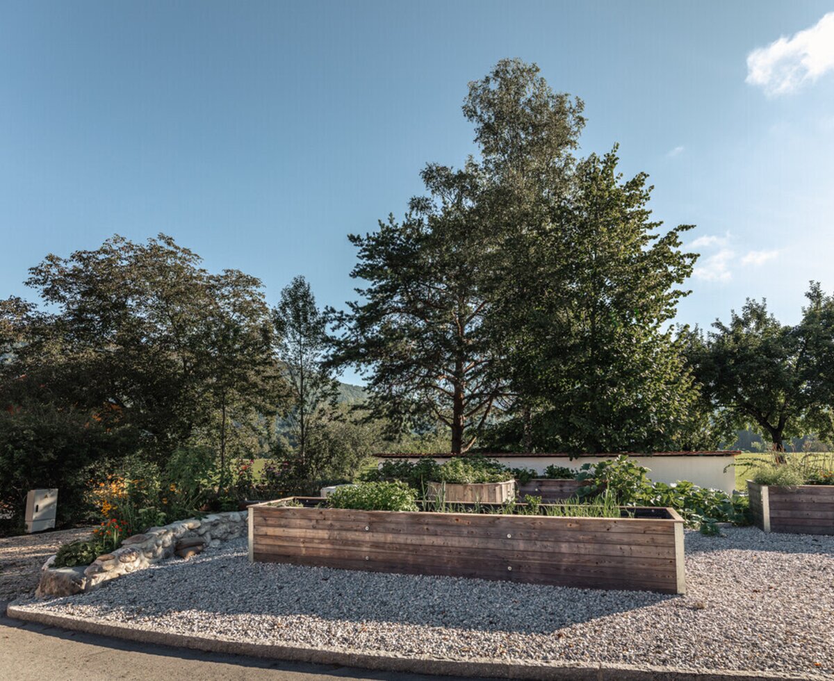The farmhouse grounds feature wooden raised garden beds on a gravel surface, surrounded by mature trees and distant hills.