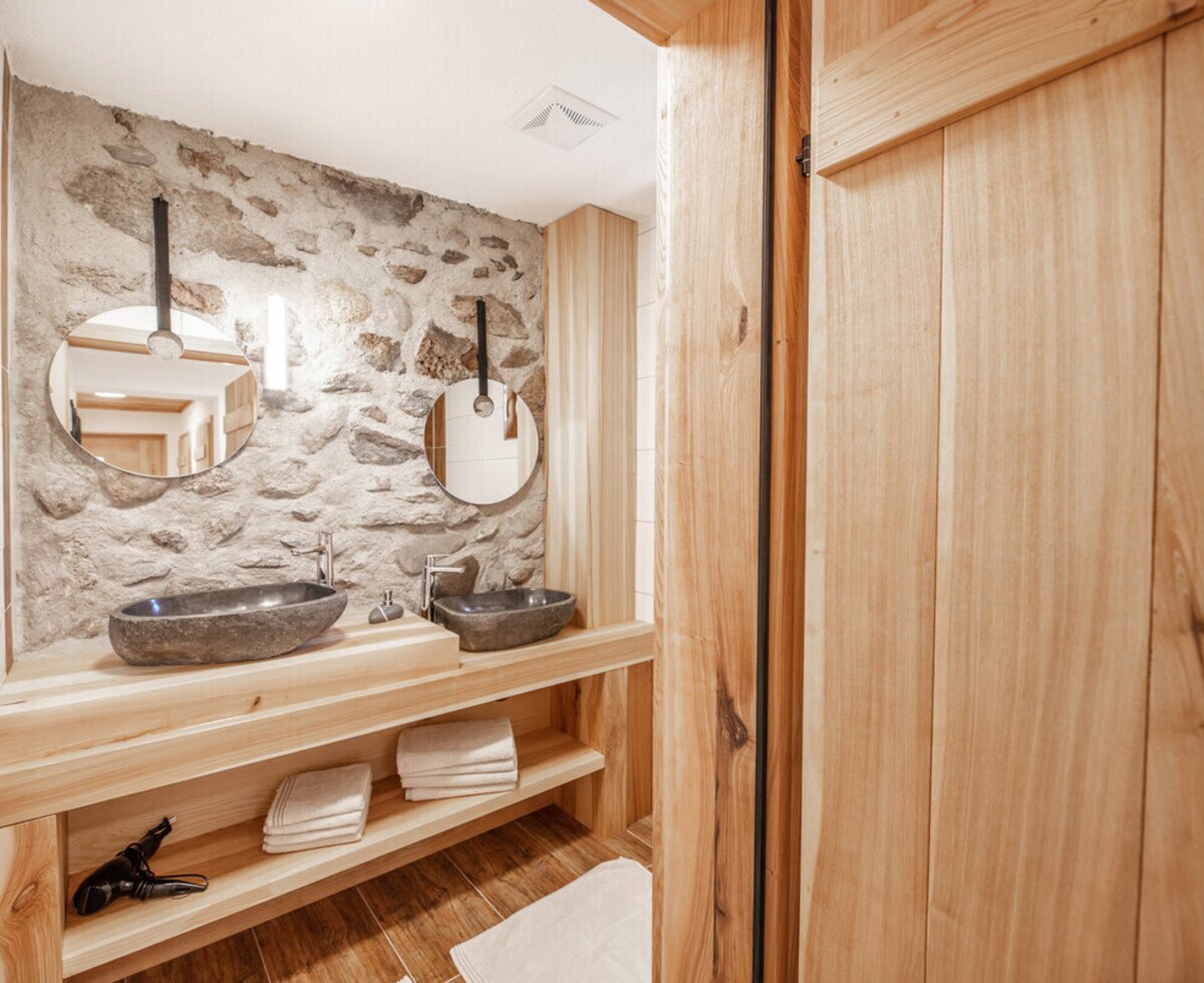 The farm house bathroom features two stone vessel sinks on a wooden vanity with open shelving, round mirrors, and a stone-clad wall.