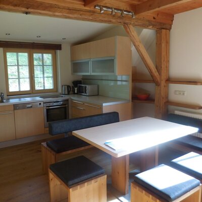The fully equipped kitchen and dining area of the holiday house, featuring a dishwasher, oven, microwave, coffee maker, and a dining table with benches, with a television mounted on the wall.