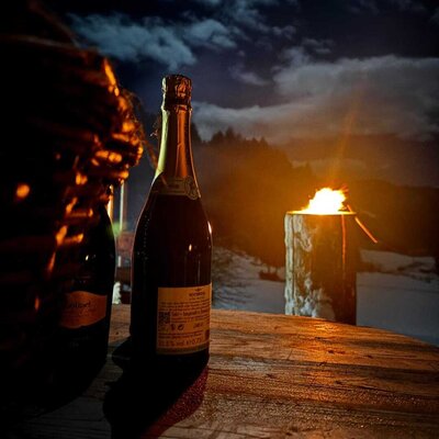 A bottle of sparkling wine on a rustic wooden table, illuminated by a warm Swedish torch at night on the farmhouse.