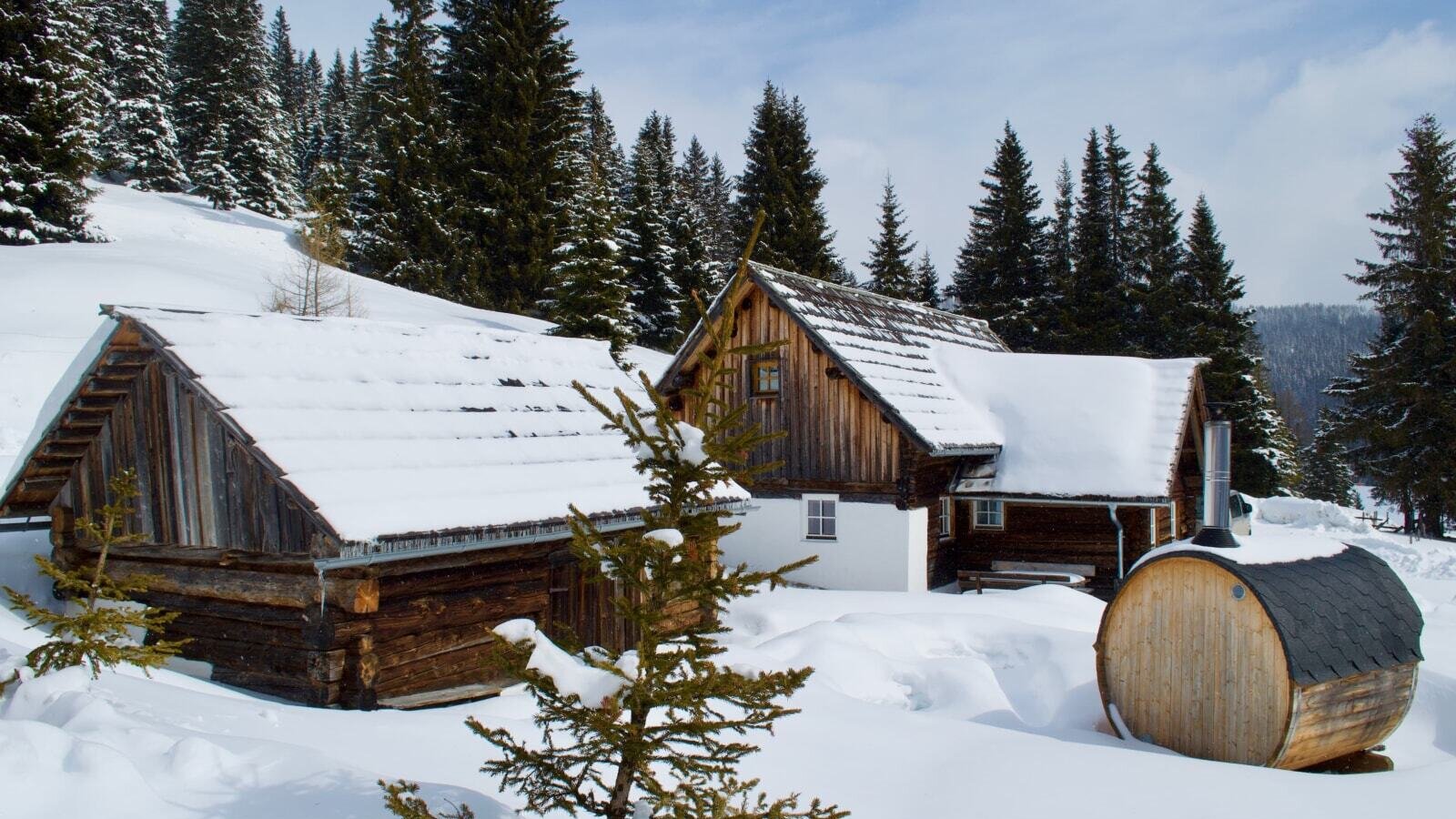 The exterior of the farmhouse in a snowy mountain setting, featuring a barrel-shaped outdoor structure with a chimney and surrounding pine trees.