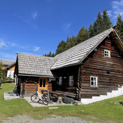 The exterior of the wooden farmhouse features a shingled roof, an outdoor seating area, and a barrel-shaped structure on the grassy grounds, backed by a forest.