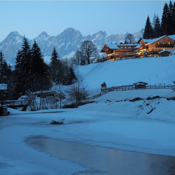 The illuminated farmhouse is set on a snowy hillside with pine trees and mountains in the background, overlooking a partially frozen stream in the foreground.
