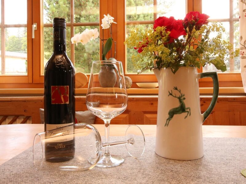 Table in the farmhouse with a wine bottle, glasses, and a flower bouquet in front of a window with a view of greenery. (c) Hans-Peter Steiner